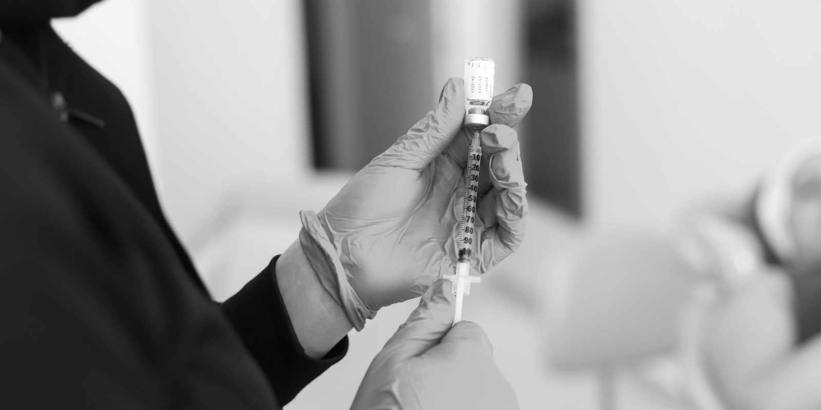 Black-and-white close-up of gloved hands drawing a clear injectable from a small glass vial into a syringe