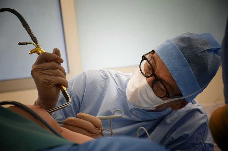 Masked surgeon in a blue cap and glasses holding a liposuction cannula attached to suction tubing