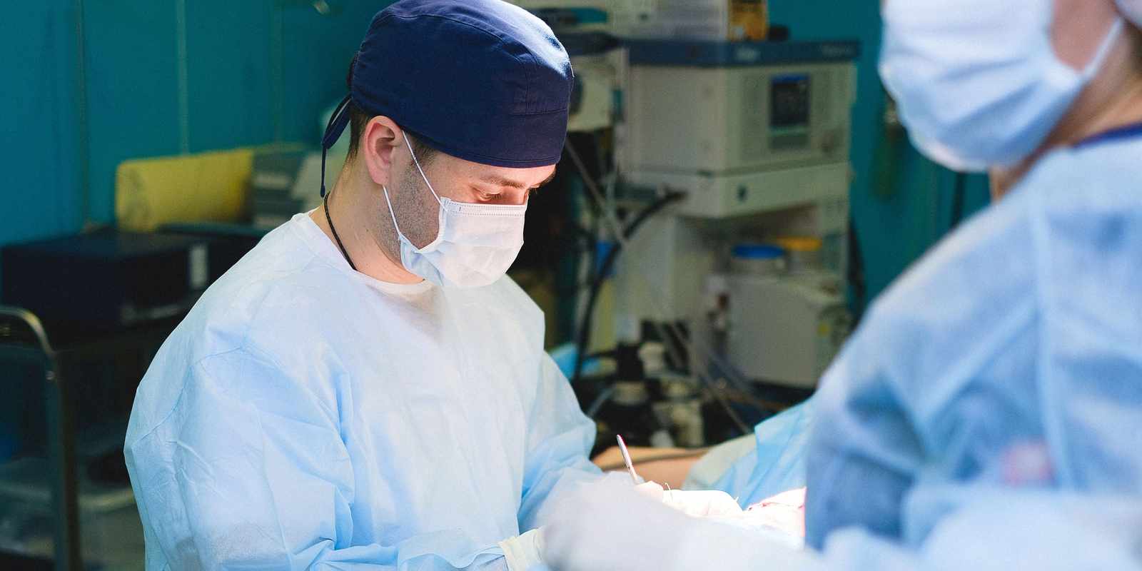 Masked surgeon in a navy cap and light-blue gown concentrating during an operation, a colleague blurred in the foreground