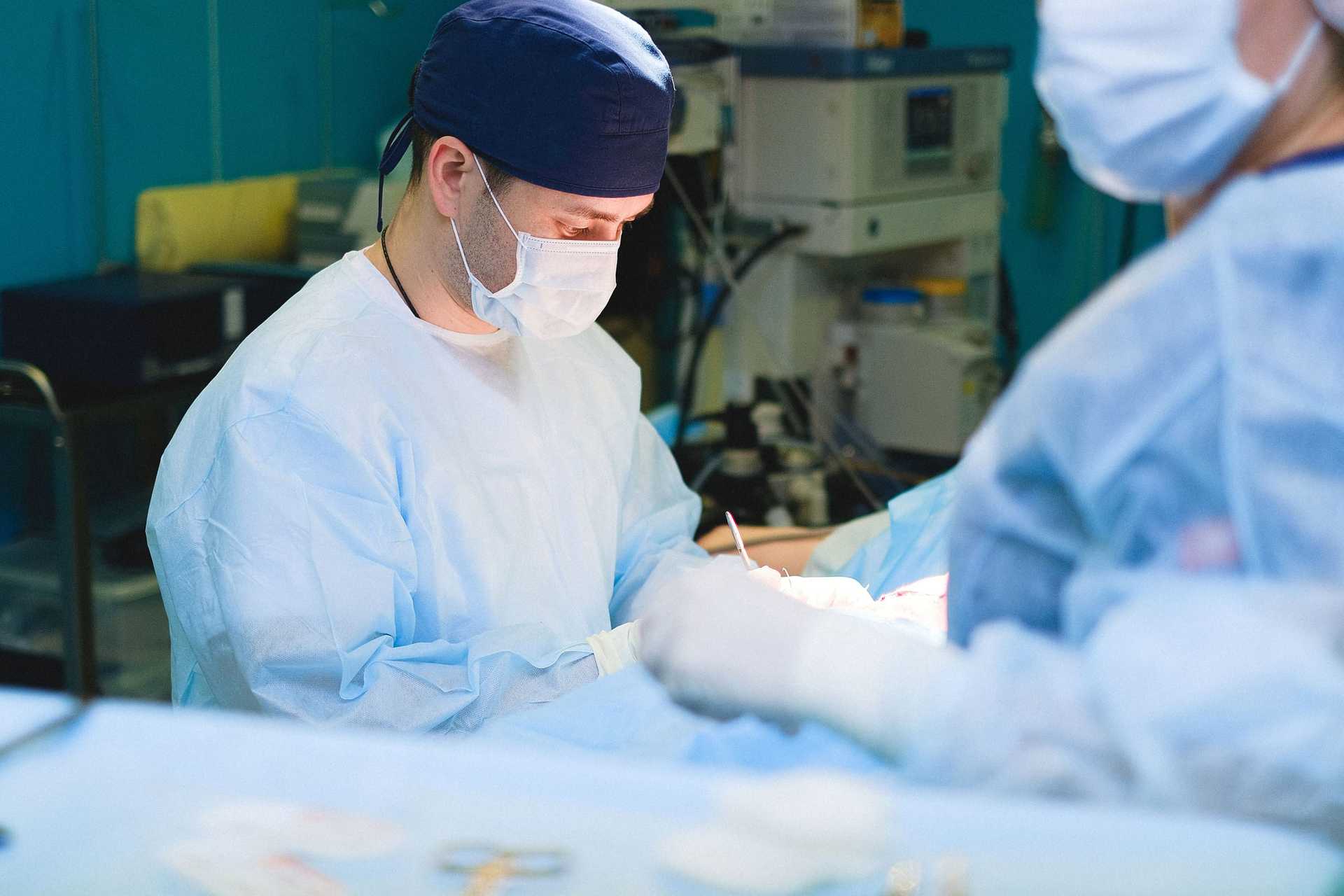 Masked surgeon in a navy cap and light-blue gown concentrating during an operation, a colleague blurred in the foreground